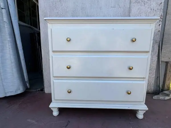 White wood 3-drawer dresser with gold round knobs and turned bun feet.